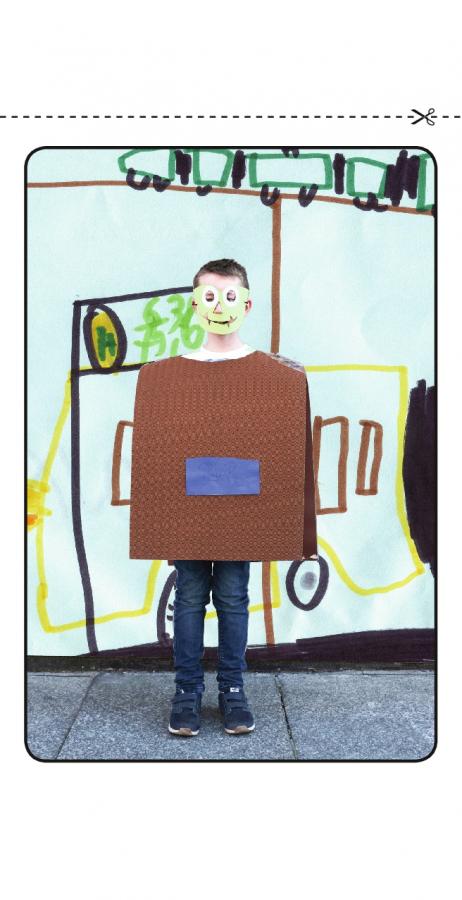 Ein Kind trägt eine Papiermaske und ein aus Karton gefertigtes quadratisches Kostüm, hinter ihm eine Wand mit Zeichnungen.