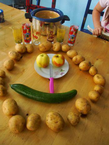 Kartoffeln in einem Kreis auf einem Tisch angeordnet, gestaltet wie ein Gesicht: Kinder haben eine Gurke als Mund, ein Küchenmesser als Nase, einen Teller mit zwei Äpfeln als Augen, Trinkbecher als Haare und dahinter einen Topf platziert.