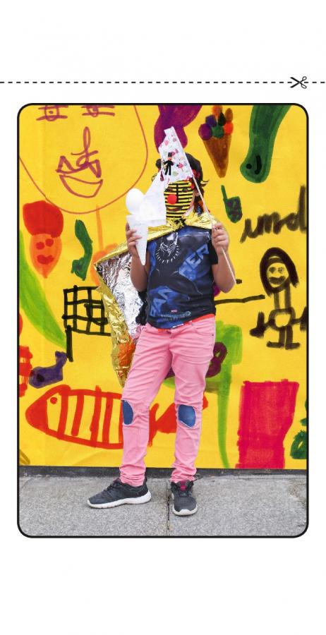 Ein kleines Mädchen trägt einen Umhang, hält eine kleine bunte Flagge in der Hand und steht vor einer Wand, die mit verschiedenen Farben bemalt ist.