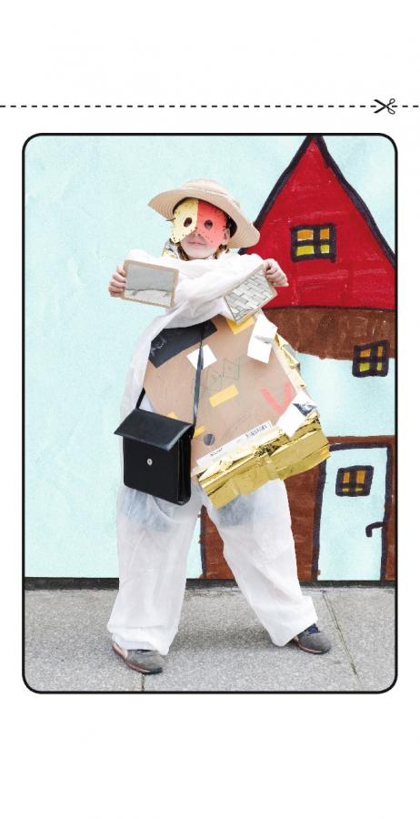 Ein kleines Mädchen trägt Kleidung mit Papierformen, hält eine Handtasche, trägt in jeder Hand einen Spiegel, sowie einen Hut und eine Papiermaske; hinter ihr eine Wand mit einem Haus gemalt