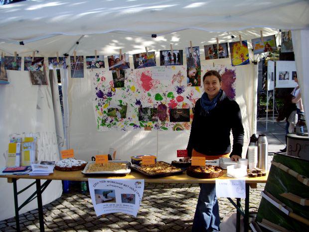 Eine Frau steht in einem weißen Zelt vor einem Tisch mit Süßigkeiten, über ihrem Kopf hängt eine Schnur mit Fotos, hinter ihr ist die Zeltwand mit Kinderzeichnungen dekoriert