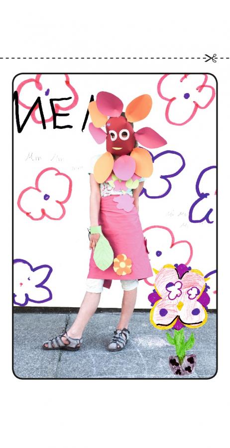 Ein kleines Mädchen steht und trägt eine Papiermaske in Blumenform, hinter ihr eine Wand mit Blumenmalereien.