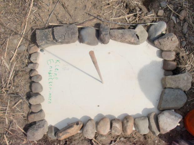 Steine auf einem Erdgrund in rechteckiger Anordnung, in der Mitte ein weißes Blatt mit kleinem Text und ein Holzstab in der Mitte gesteckt
