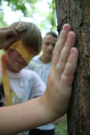 Ein Kind legt die Hand auf einen Baum, daneben ein anderes Kind mit verbundenen Augen, das erste Kind versucht das gelbe Tuch mit der anderen Hand von den Augen seines Freundes zu nehmen, hinter ihnen steht ein weiterer Junge.