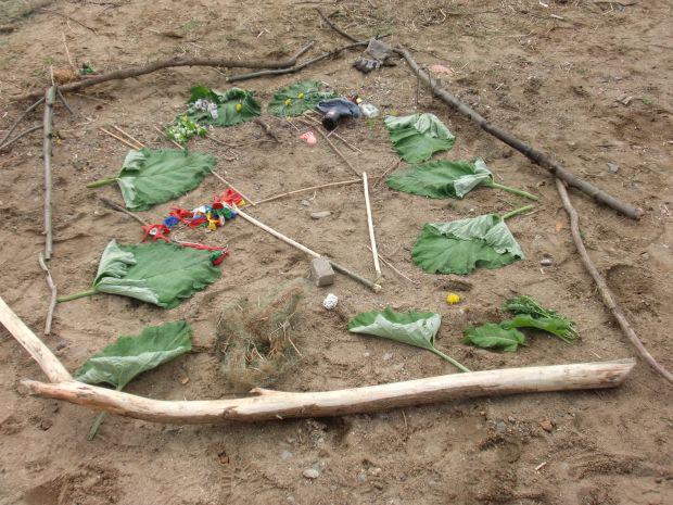 Ein Quadrat aus Holzstäbchen auf einem Erdgrund, darin Blätter, und in der Mitte ein kleines Dreieck aus kleineren Stäbchen.