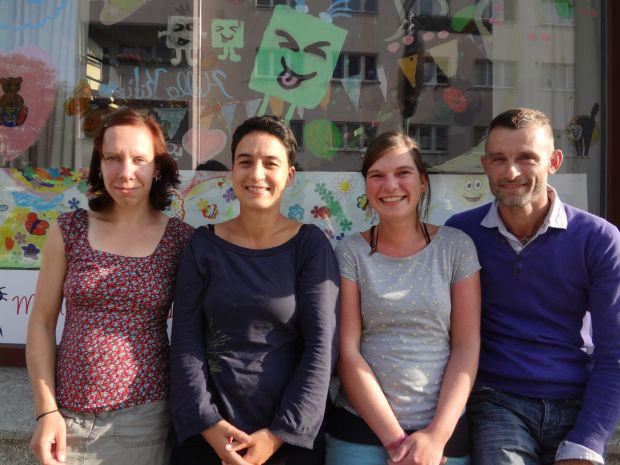 Drei Frauen und ein Mann lächeln in die Kamera, sie stehen vor einer Glasfassade, die mit Kinderzeichnungen dekoriert ist.