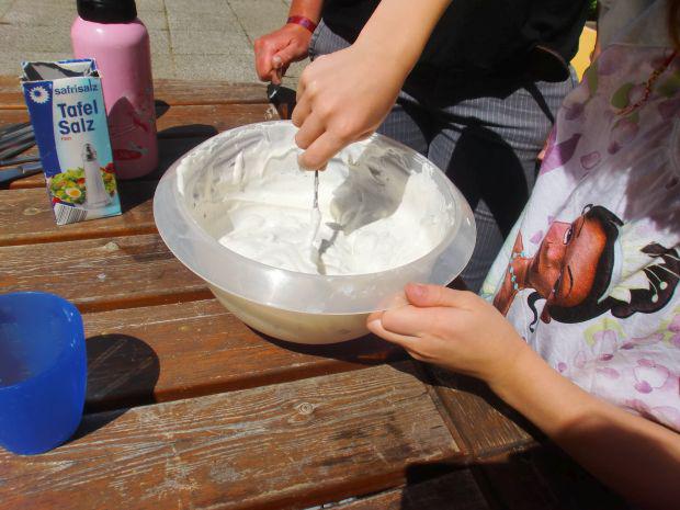 Ein Kind mischt Gips mit Wasser in einer weißen Schüssel auf einem Holztisch, neben ihm steht sein Freund.