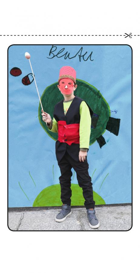 Ein Kind trägt einen rosa Fez und eine rosa Papiermaske, hinter ihm eine Wand mit Zeichnungen.