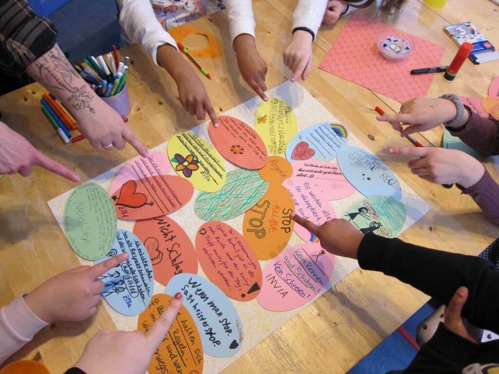 Zehn Personen um einen Tisch, nur ihre Arme sind sichtbar, sie zeigen mit den Fingern auf eine Sammlung von Papieraufklebern, die von Kindern beschriftet wurden.