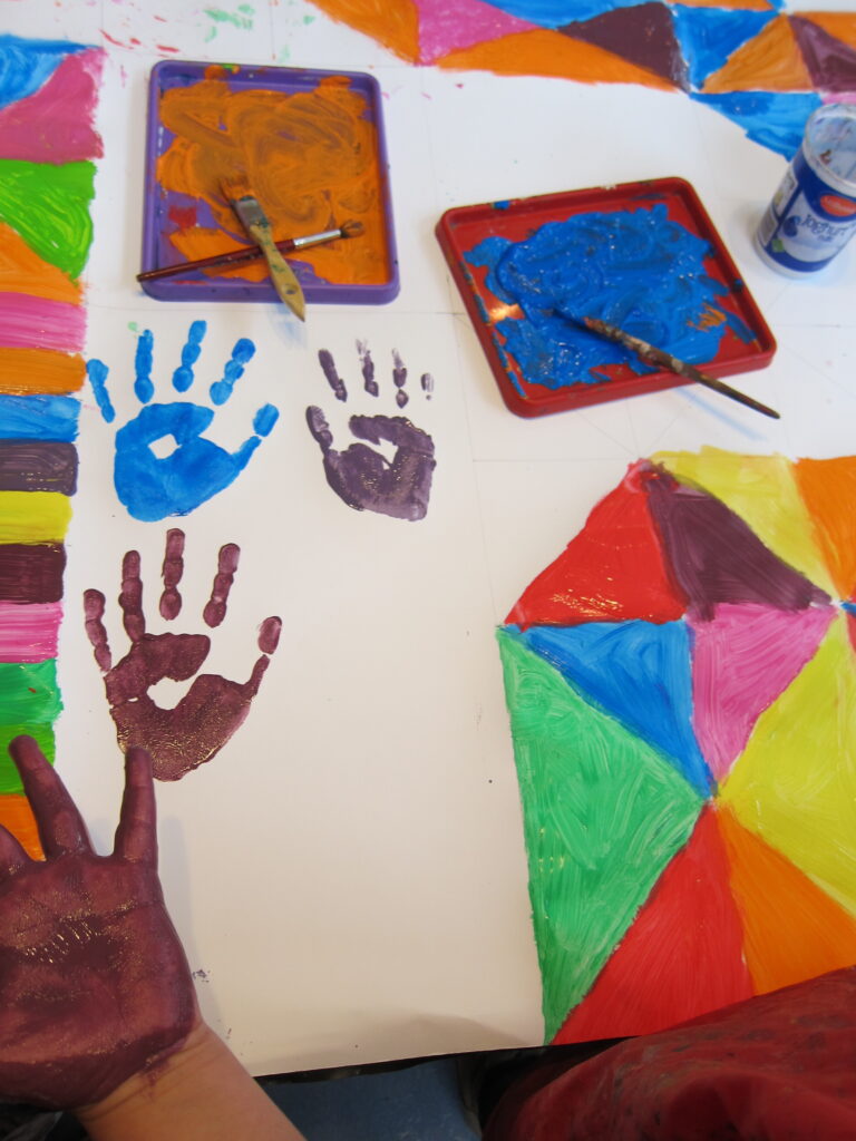 Eine Kinderhand mit Farben, davor ein Blatt Papier mit bunten Farben und einem Handabdruck des Kindes.