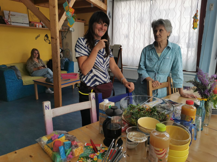 Zwei Frauen stehen vor einem Tisch mit Salat und Getränken, hinter ihnen sitzt eine weitere Frau auf einem Sofa
